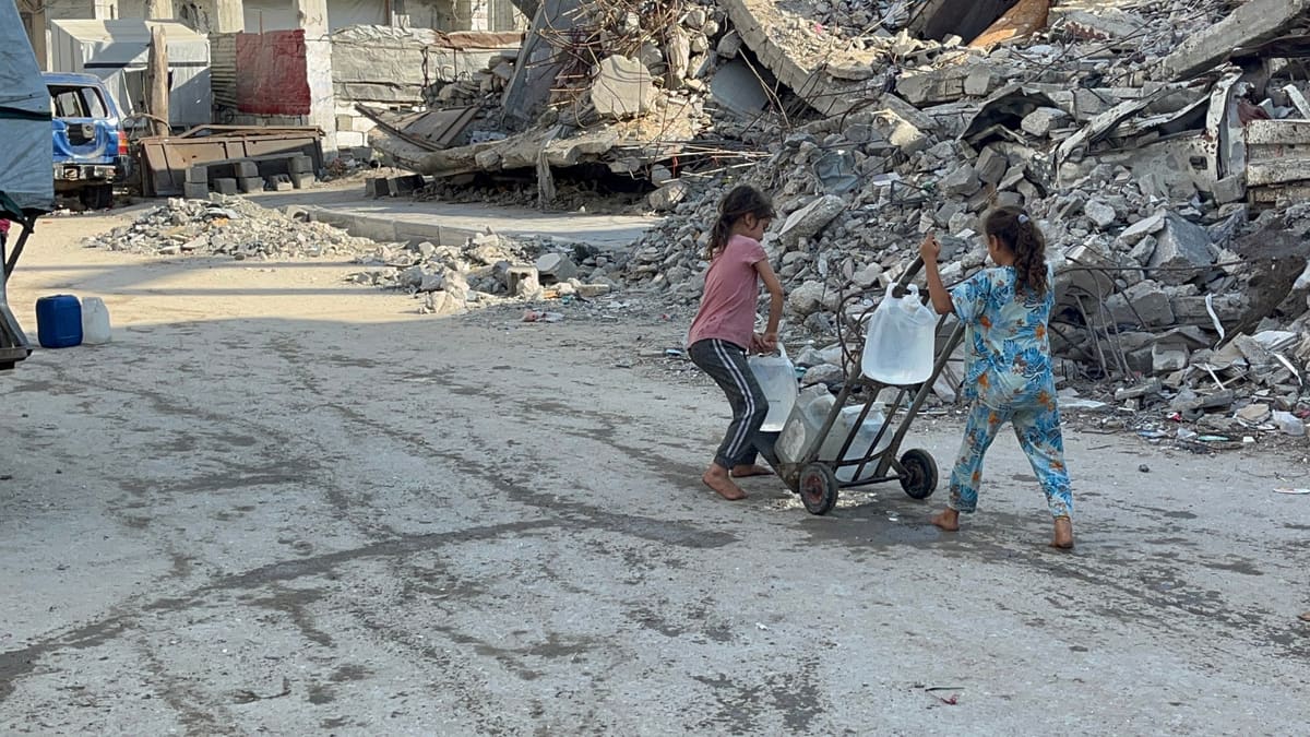 Dua anak membawa jeriken air dengan troli di jalan yang dipenuhi reruntuhan